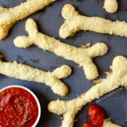 Top-down view of Cheesy Halloween Breadstick Bones next to a small bowl containing marinara sauce.