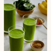 Three glasses filled with Healthy Green Smoothie mix next to a bowl containing fresh spinach and a smaller bowl containing ground flaxseed.