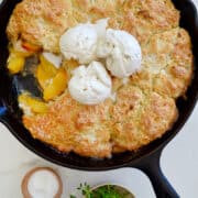 Peach Cobbler with buttermilk biscuits in a cast-iron skillet topped with three scoops of vanilla bean ice cream.