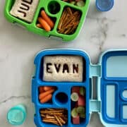 A top-down view of two Bentgo lunchboxes containing sandwiches with the names "Julian" and "Evan" cut out from cookie cutters, baby carrots, pretzel sticks and fresh fruit.