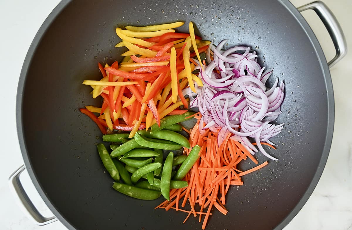 Shredded carrots, snap peas, sliced bell peppers and sliced red onions in a wok.
