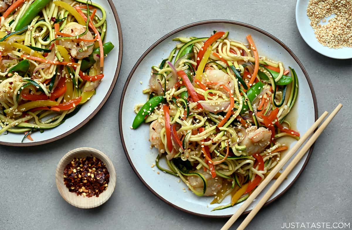 Two dinner plates with chopsticks resting on the edge containing zucchini noodle stir-fry with shrimp.