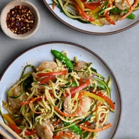 Zucchini noodle stir-fry shrimp on a dinner plate next to a small bowl containing crushed red pepper flakes.