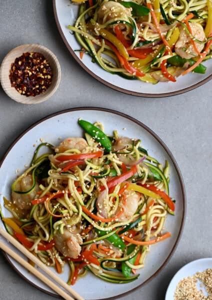 Zucchini noodle stir-fry shrimp on a dinner plate next to a small bowl containing crushed red pepper flakes.