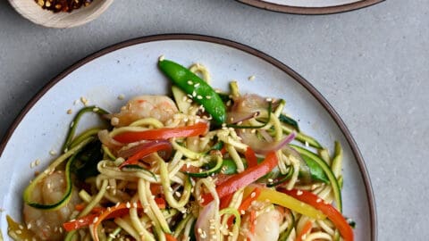 Zucchini noodle stir-fry shrimp on a dinner plate next to a small bowl containing crushed red pepper flakes.
