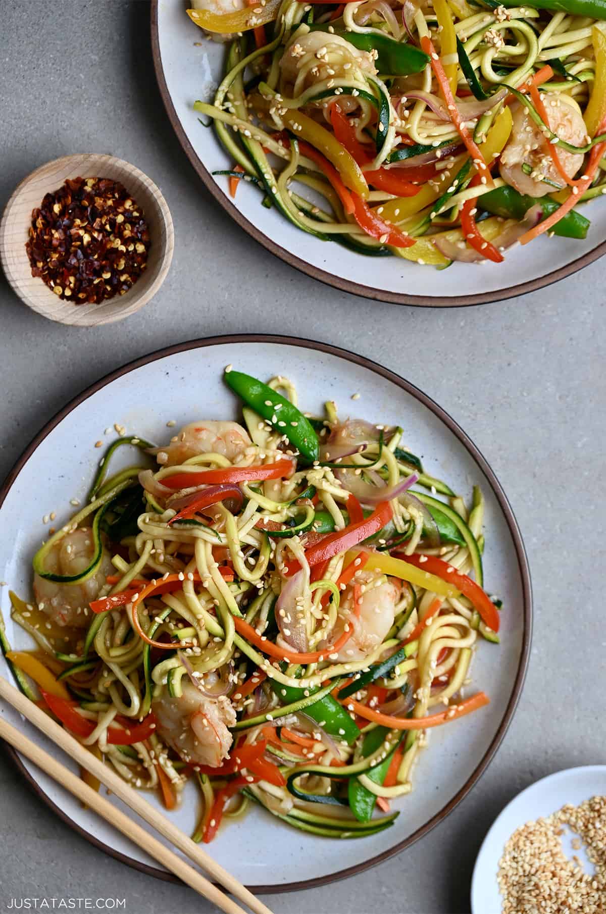 Zucchini noodle stir-fry shrimp on a dinner plate next to a small bowl containing crushed red pepper flakes.