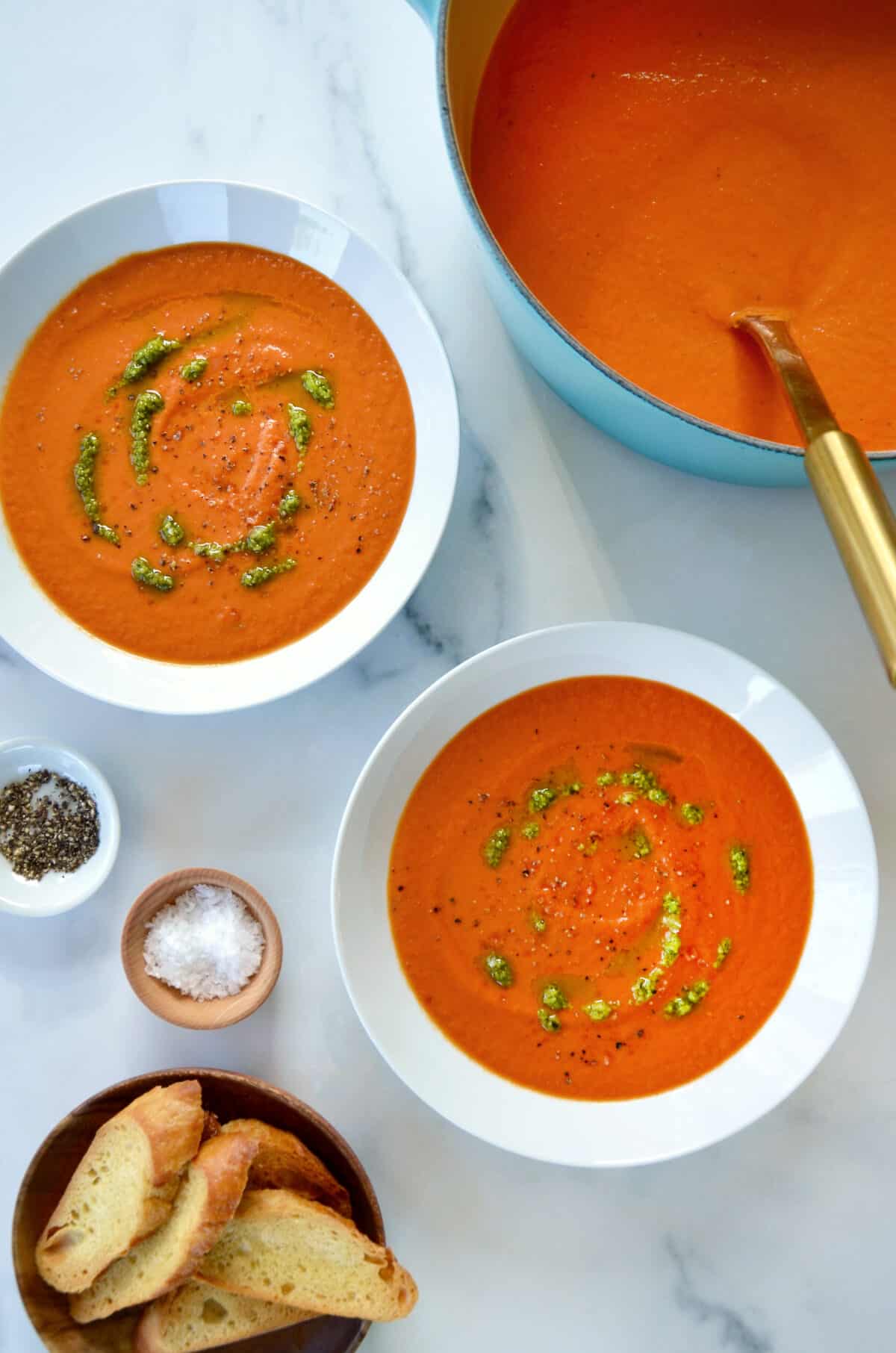 Two white bowls are filled with tomato soup and drizzled with basil pesto. Small bowls of salt, pepper and sliced baguette are nearby.
