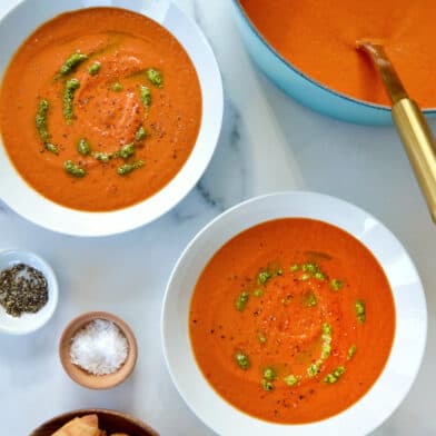 Two white bowls are filled with tomato soup and drizzled with basil pesto. Small bowls of salt, pepper and sliced baguette are nearby.