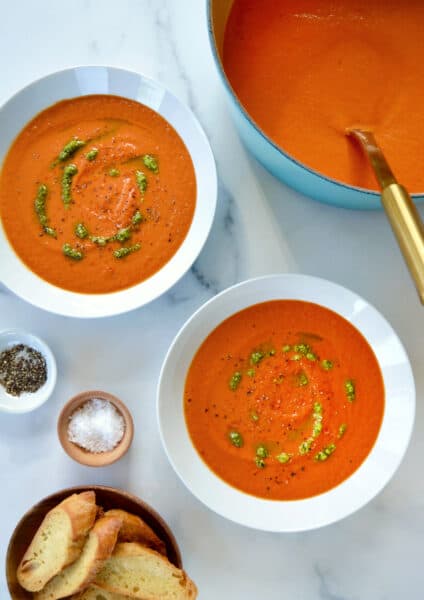 Two white bowls are filled with tomato soup and drizzled with basil pesto. Small bowls of salt, pepper and sliced baguette are nearby.