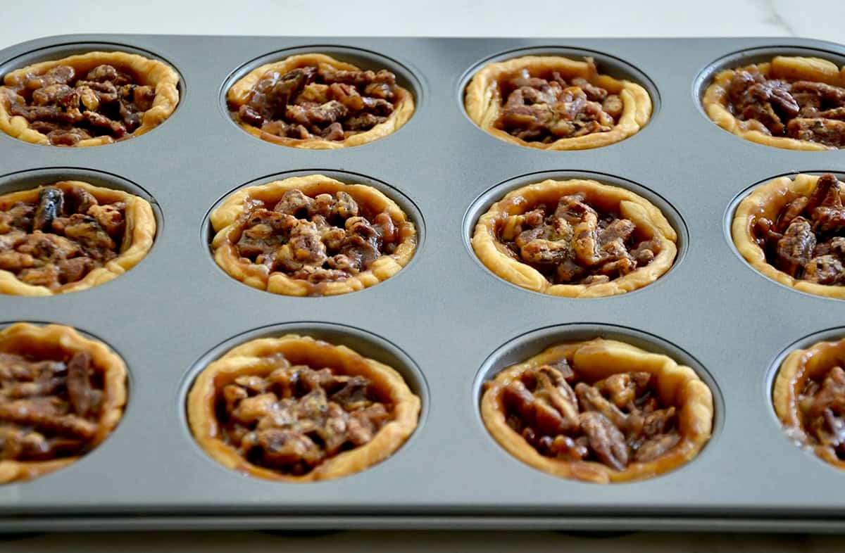 Freshly baked golden brown individual pecan pies in a muffin pan.