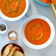 Two white bowls containing Creamy Homemade Tomato Soup drizzled with basil pesto next to a stockpot containing soup and small bowls containing black pepper, salt, and crusty toasts.