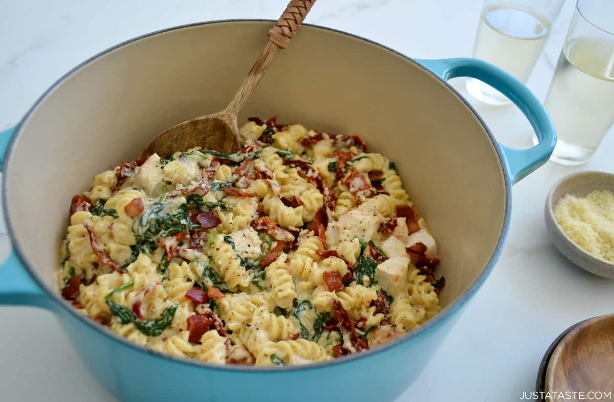 A Dutch oven with Tuscan chicken pasta with spinach and sun-dried tomatoes with a wooden spoon.