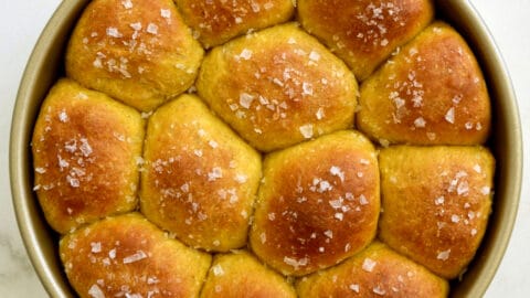 A round baking pan containing golden brown pumpkin dinner rolls garnished with large-flake sea salt.