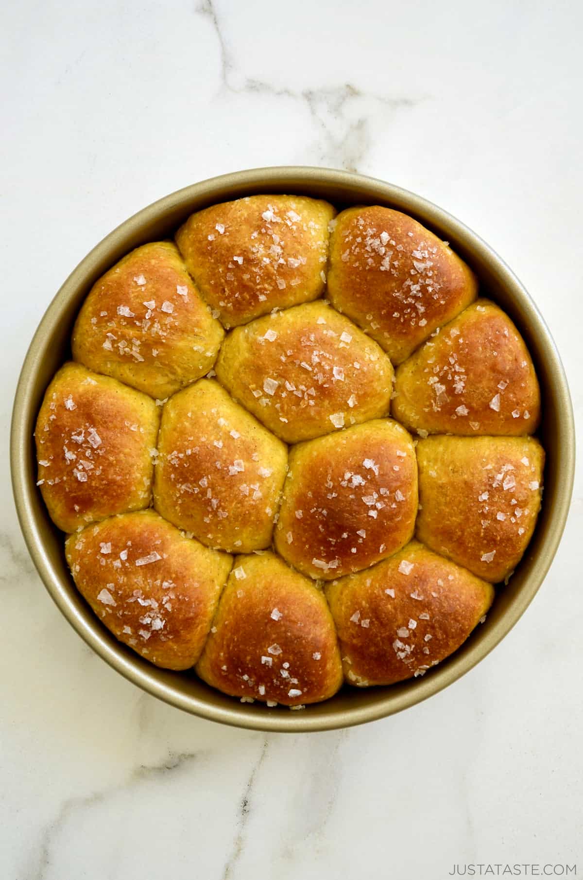 A round baking pan containing golden brown pumpkin dinner rolls garnished with large-flake sea salt.