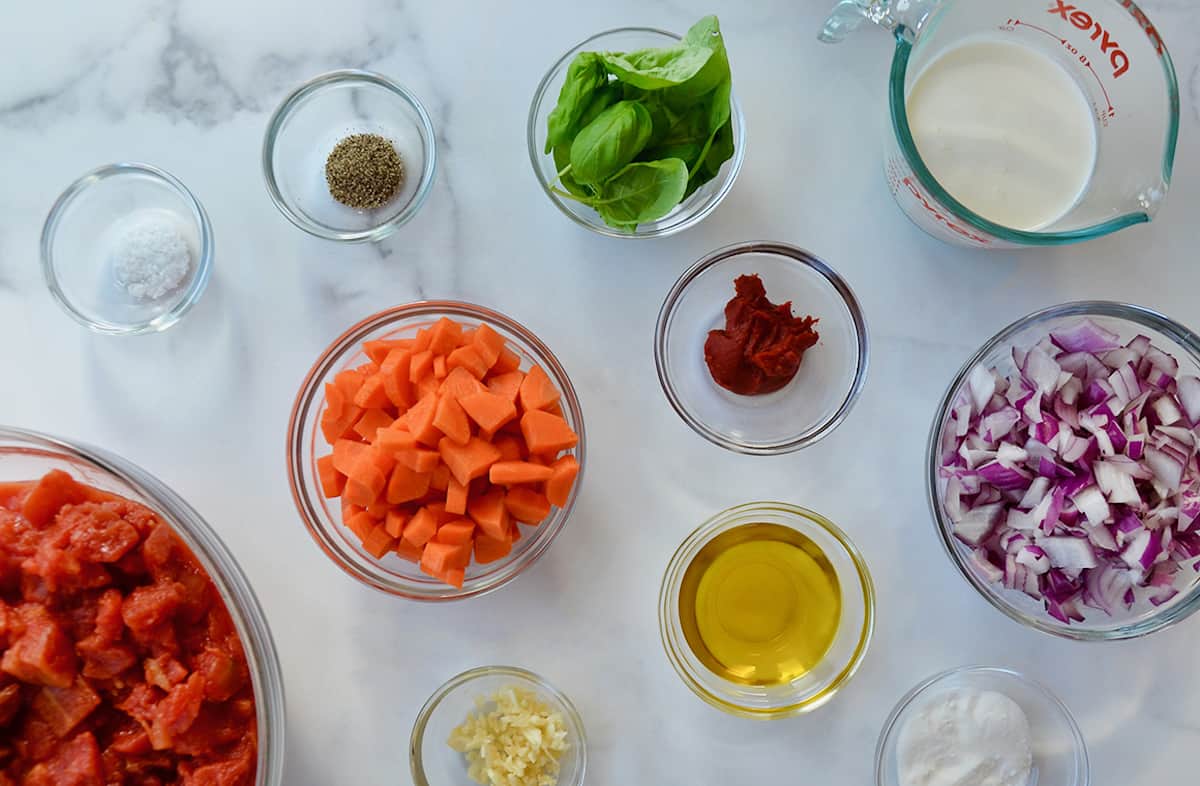 Glass bowls are filled with ingredients for creamy tomato soup, including diced carrots, olive oil, basil leaves, canned tomatoes, chopped red onions, heavy cream and seasonings.