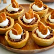 A wood plate containing Muffin Tin Mini Pecan Pies topped with whipped cream and pie crust leaves.