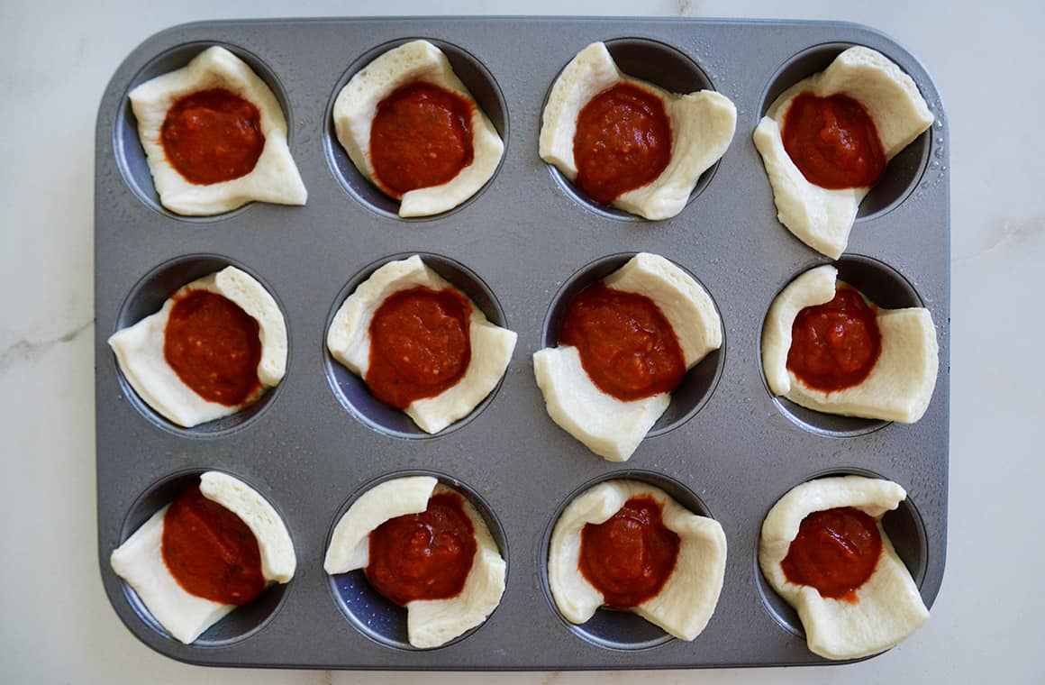 A muffin tin containing unbaked dough and marinara sauce