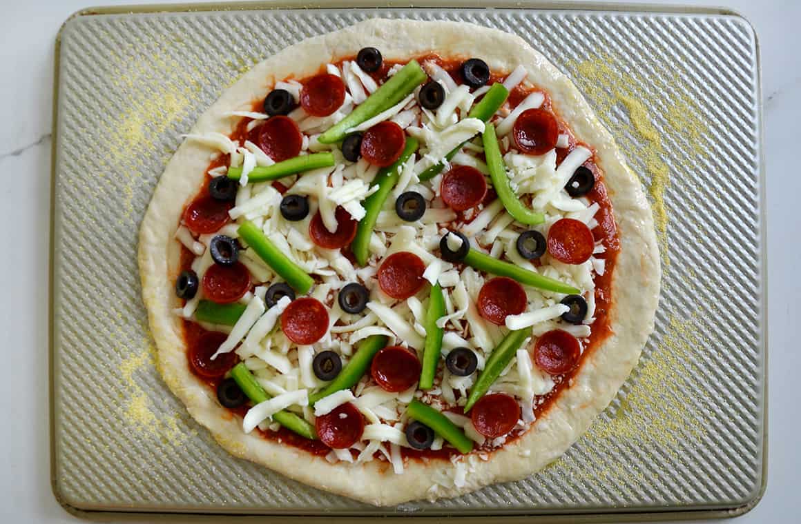 An unbaked pizza on a gold baking sheet lined with cornmeal