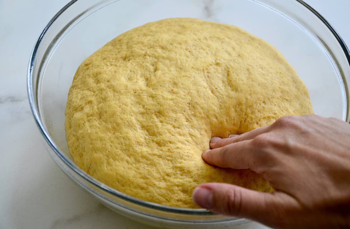 A hand pressing into a glass bowl containing proofed bread dough.