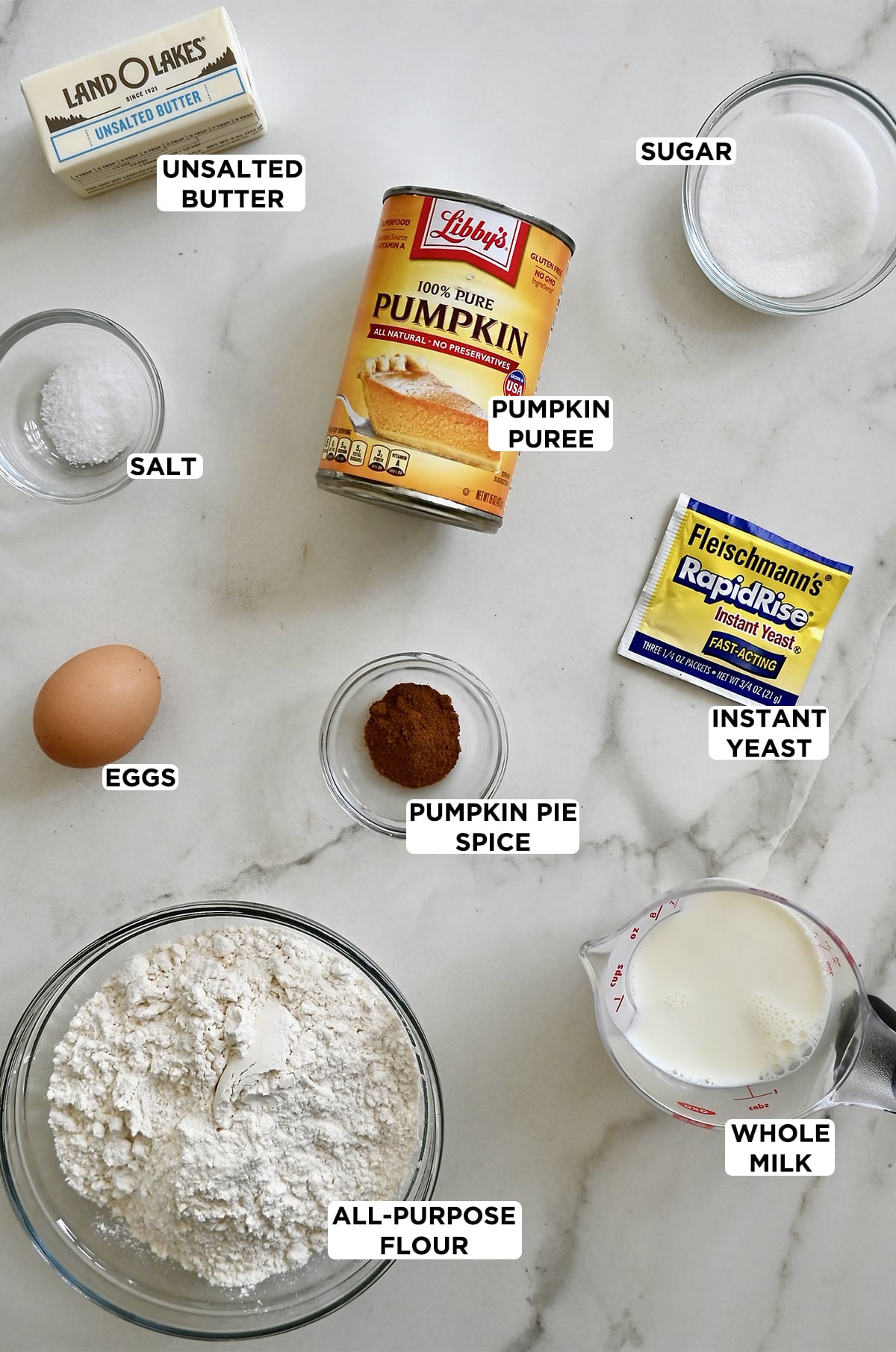 Various sizes of clear bowls containing the ingredients needed to making pumpkin dinner rolls, including sugar, instant yeast, flour, pumpkin pie spice, egg, salt a can of pumpkin puree and a liquid measuring cup containing milk. 