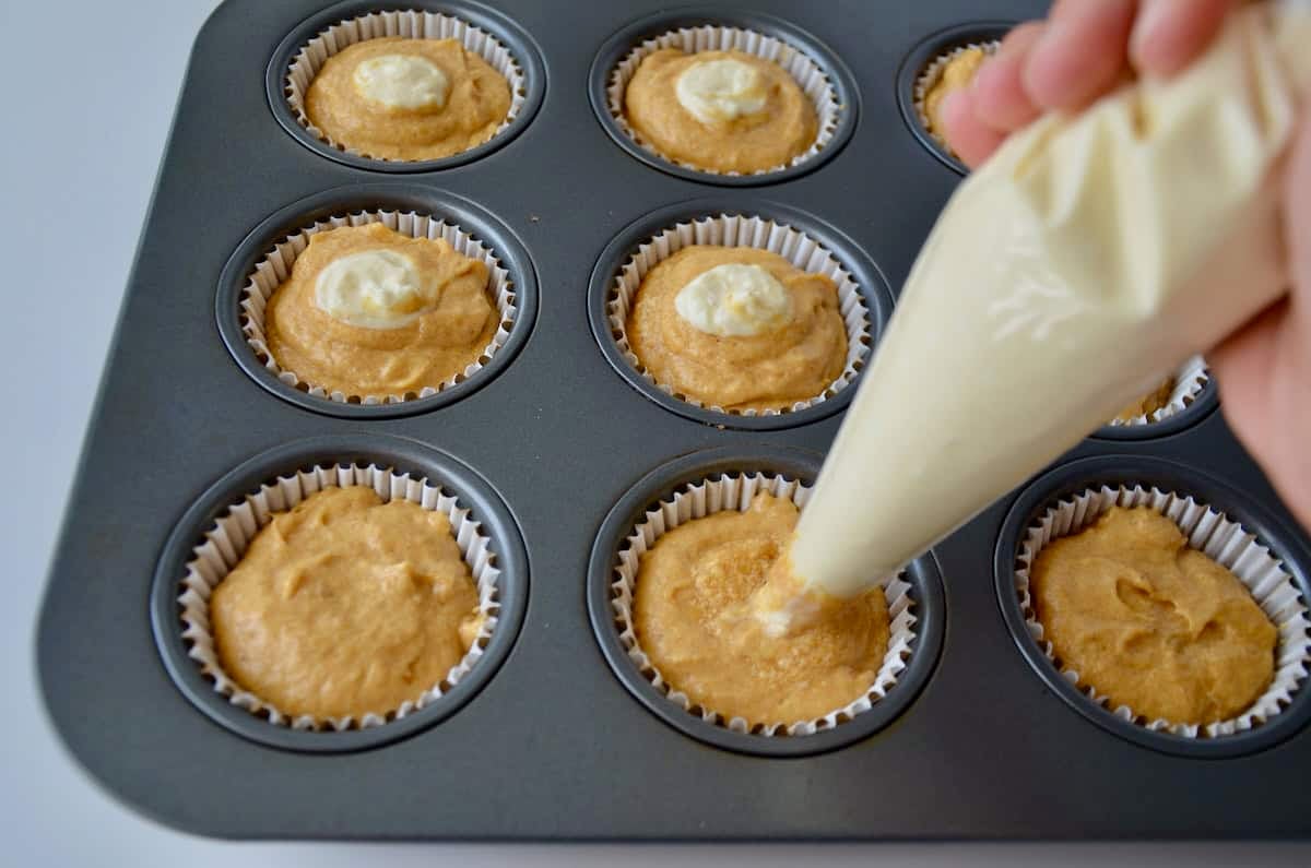 Piping a cheesecake mixture from a plastic piping bag into the center of pumpkin muffin batter.