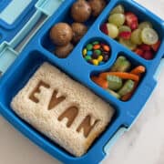 A Bentgo lunch box containing a sandwich with cutout letters that reads, "Evan", protein balls, fresh fruit, carrots and mini chocolate-covered candies.