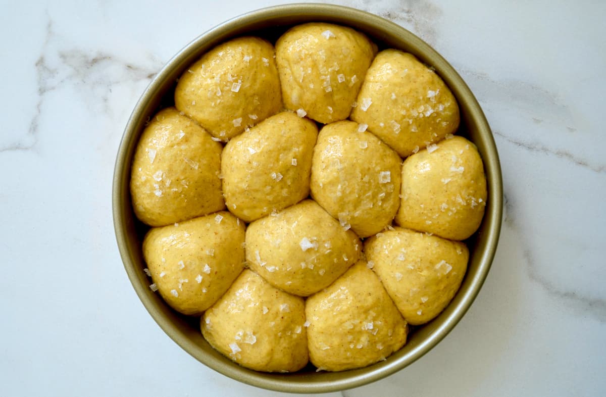 Unbaked pumpkin dinner rolls topped with large-flake sea salt in a round pan. 