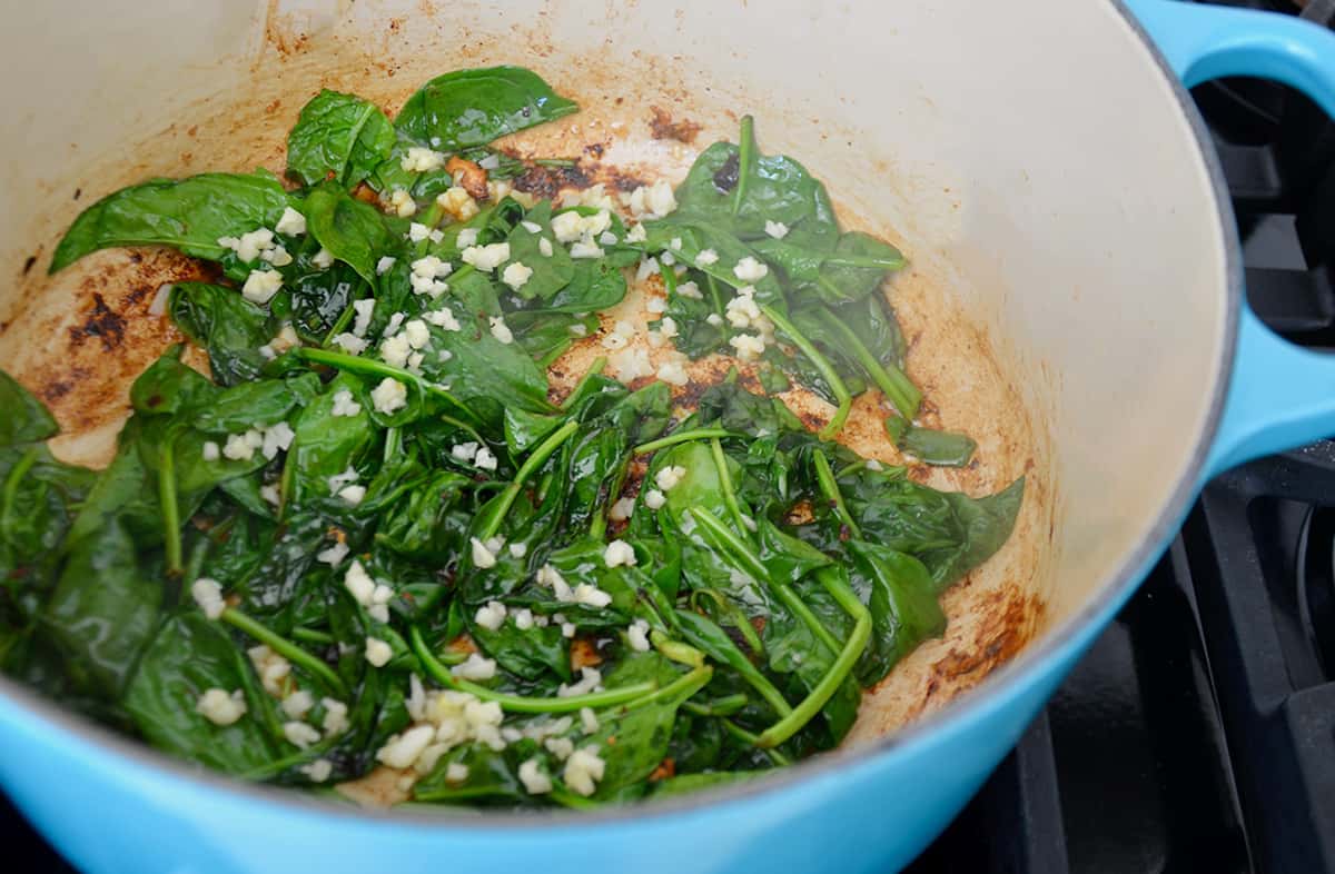 A blue Dutch oven with wilted spinach leaves and minced garlic on a stovetop.