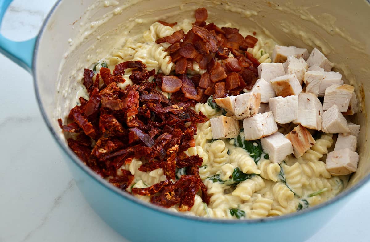 Sun-dried tomatoes, crispy bacon, diced chicken and wilted spinach over pasta in a creamy white sauce in a blue Dutch oven.