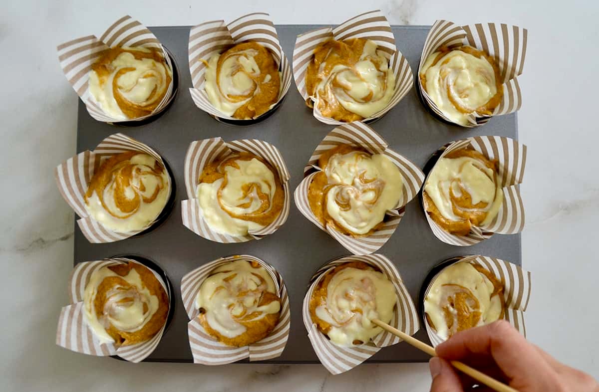 Cupcake liners filled with pumpkin batter and cheesecake-like filling are being swirled together with a toothpick.
