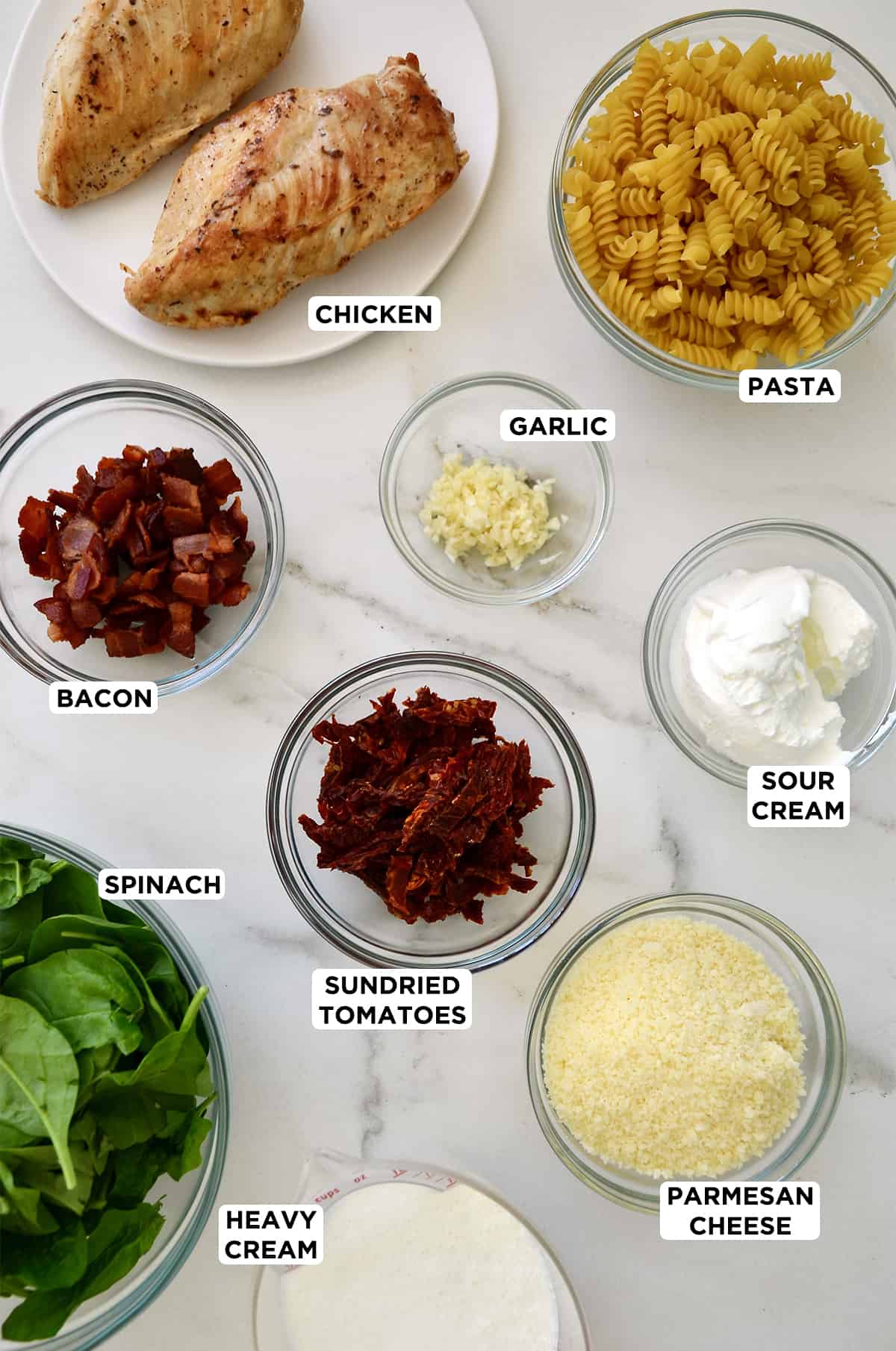 Cooked chicken breasts on a plate next to various sizes of clear bowls containing pasta noodles, minced garlic, sour cream, crispy bacon pieces, sun-dried tomatoes, grated Parmesan cheese and fresh spinach. 