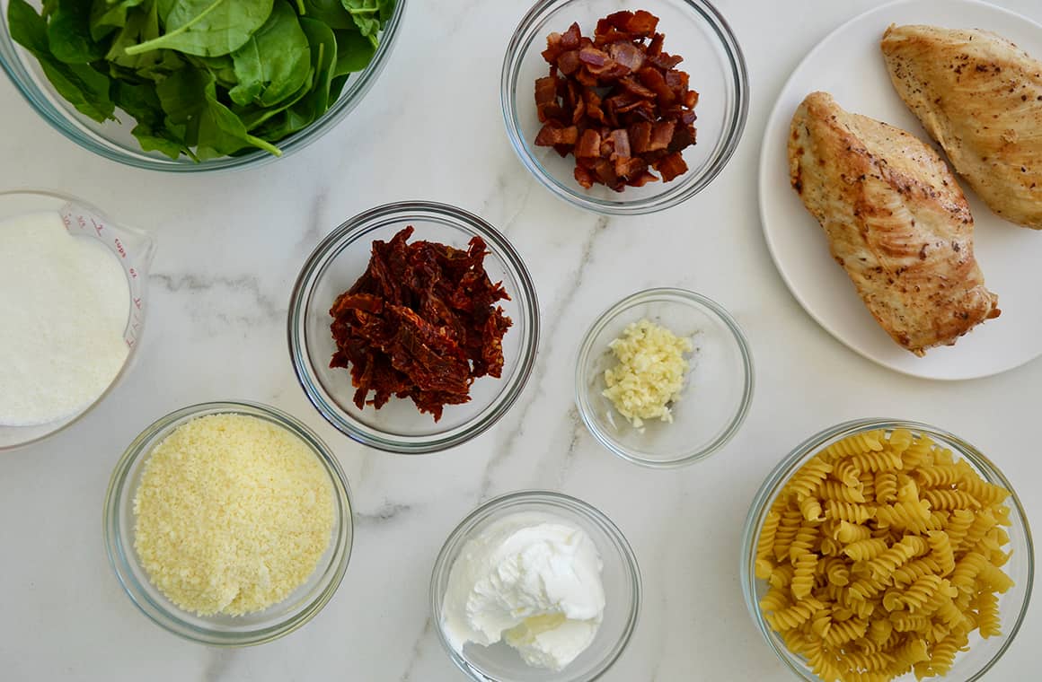 Various sizes of bowls containing spinach, sun dried tomatoes, parmesan cheese, chopped bacon, chicken breasts and minced garlic