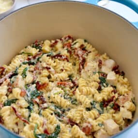 Creamy Tuscan pasta with diced chicken breasts, sun-dried tomatoes and spinach in a Dutch oven next to a small bowl containing Parmesan cheese.