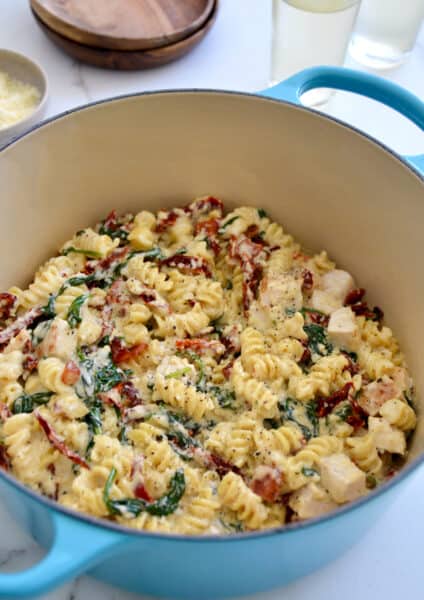 Creamy Tuscan pasta with diced chicken breasts, sun-dried tomatoes and spinach in a Dutch oven next to a small bowl containing Parmesan cheese.