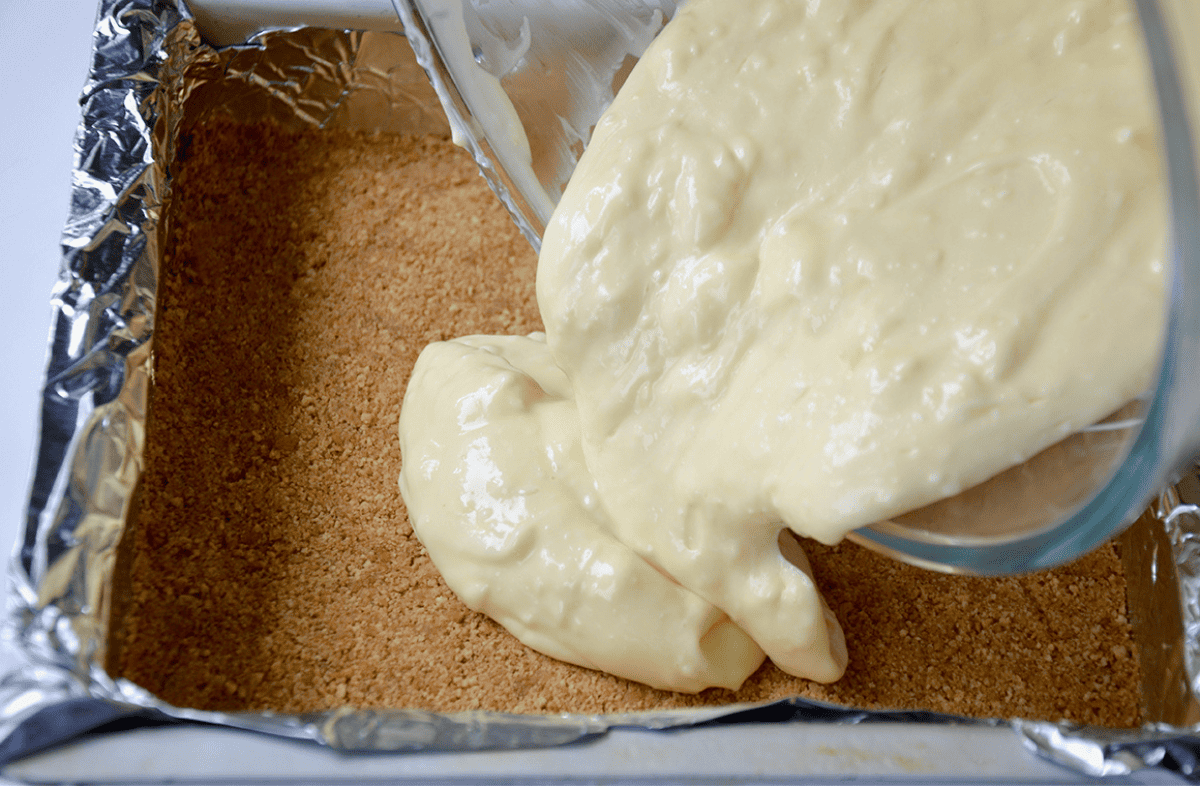 A graham-cracker crust is in a foil-lined rectangular baking pan, cheesecake filling is being poured from a clear bowl over the crust.