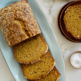 Brown speckled bananas and a dish of sanding sugar are next to a loaf of pumpkin banana bread on a blue platter.