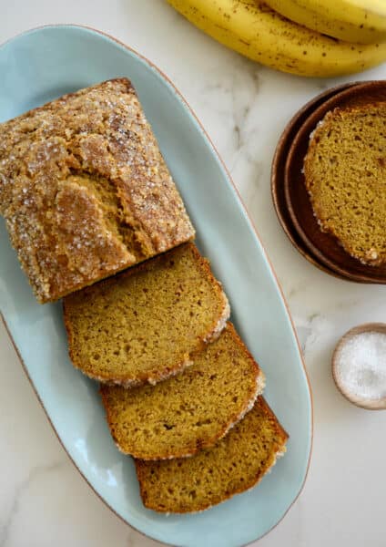 Brown speckled bananas and a dish of sanding sugar are next to a loaf of pumpkin banana bread on a blue platter.