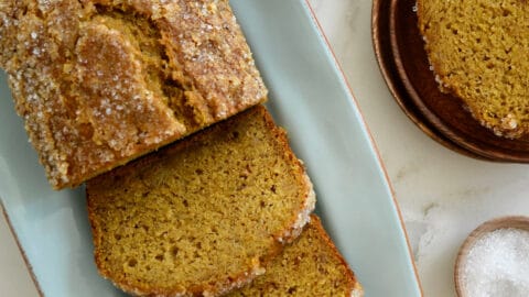 Brown speckled bananas and a dish of sanding sugar are next to a loaf of pumpkin banana bread on a blue platter.