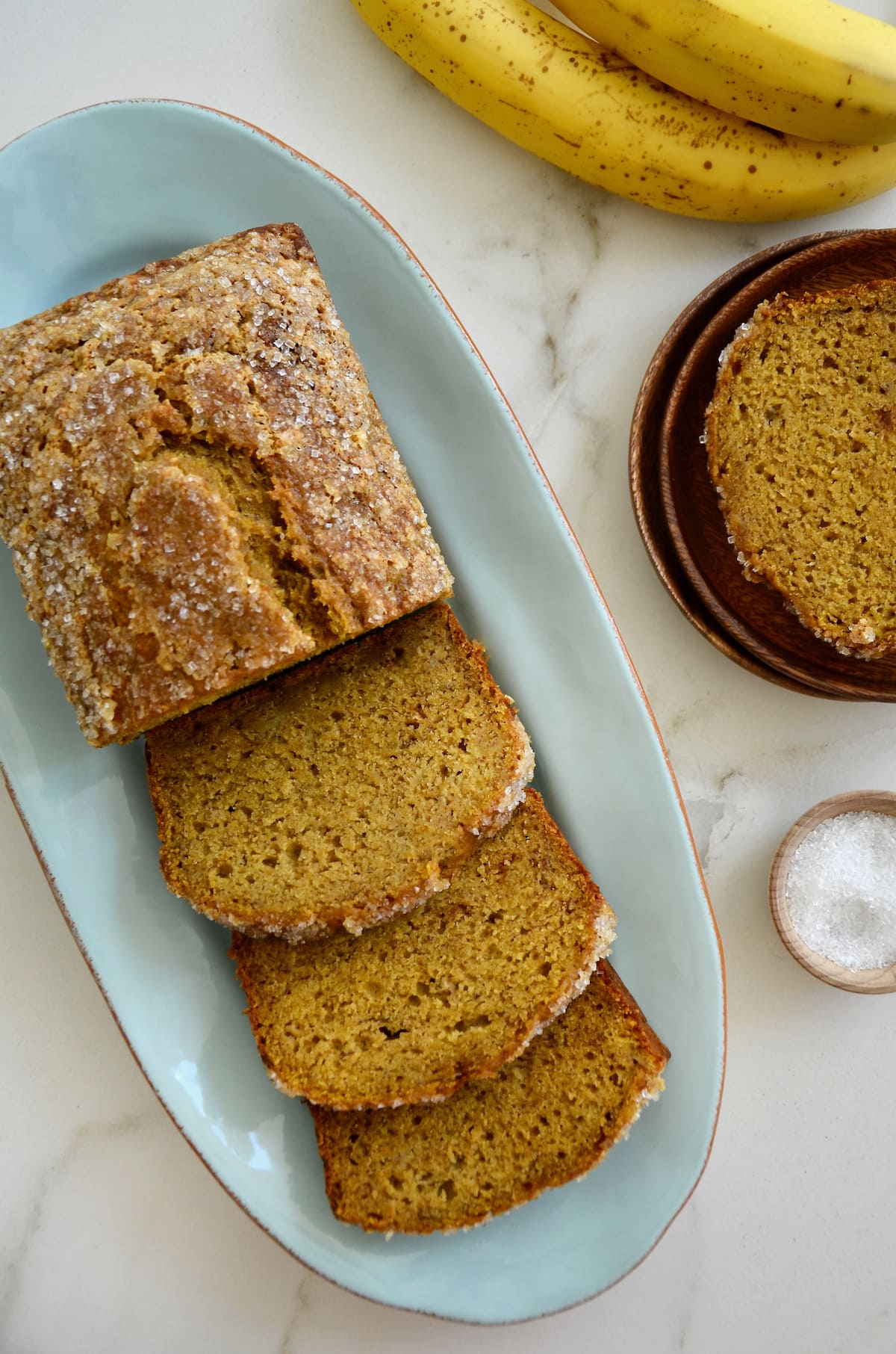 Brown speckled bananas and a dish of sanding sugar are next to a loaf of pumpkin banana bread on a blue platter.