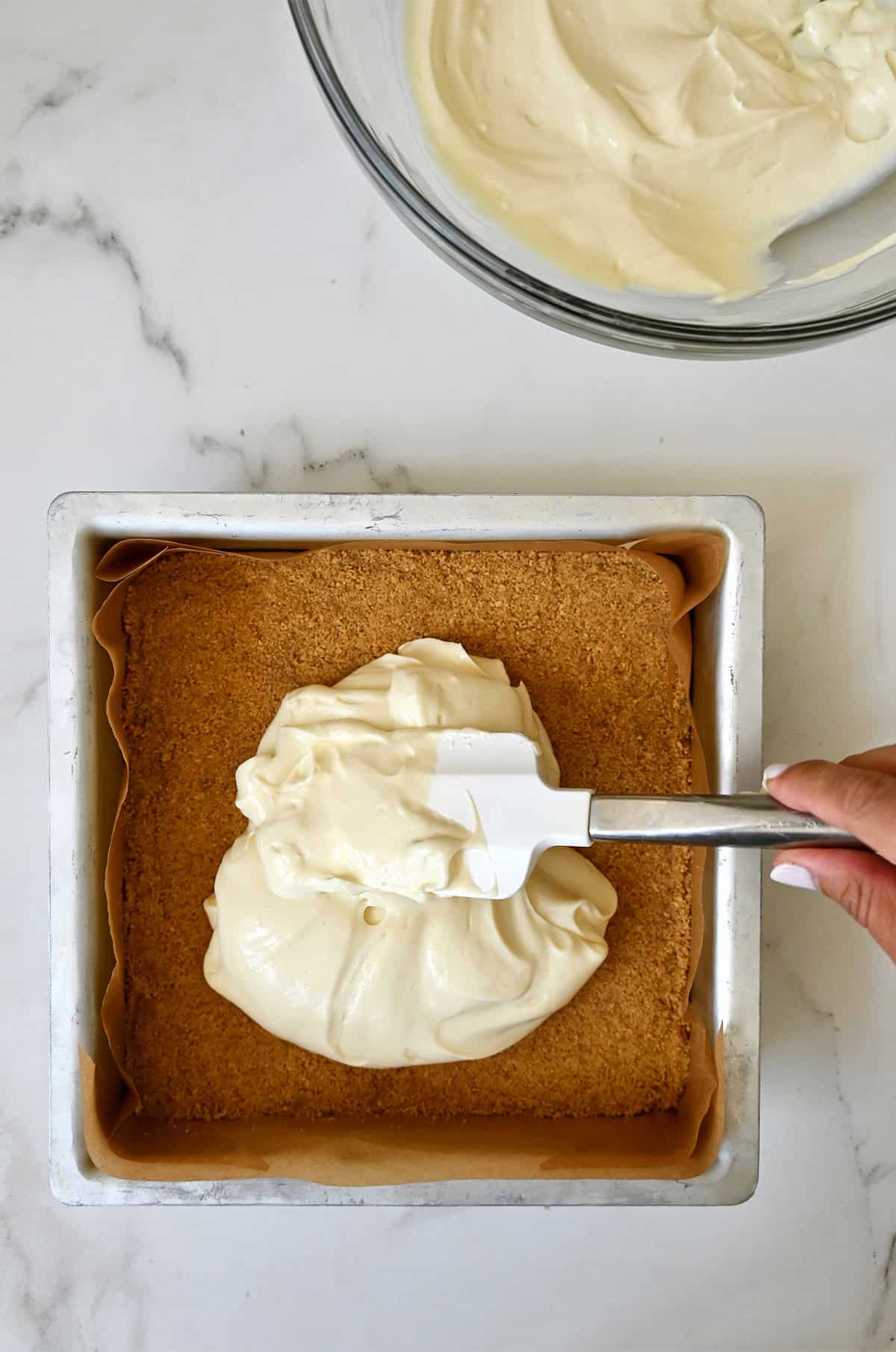 Smoothing cheesecake batter atop graham cracker crust in a square pan.