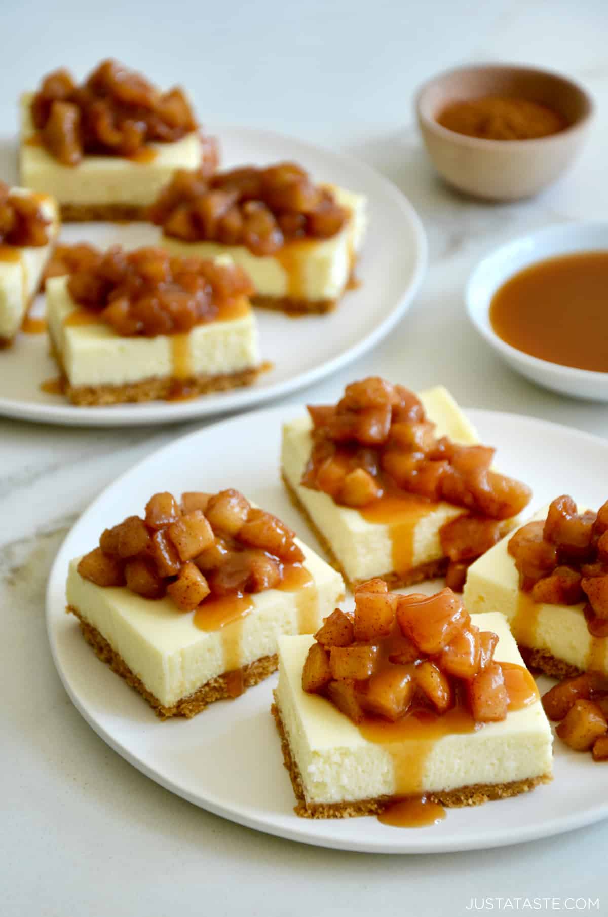 Cheesecake bars topped with sautéed apples and drizzled with caramel sauce on a white plate.