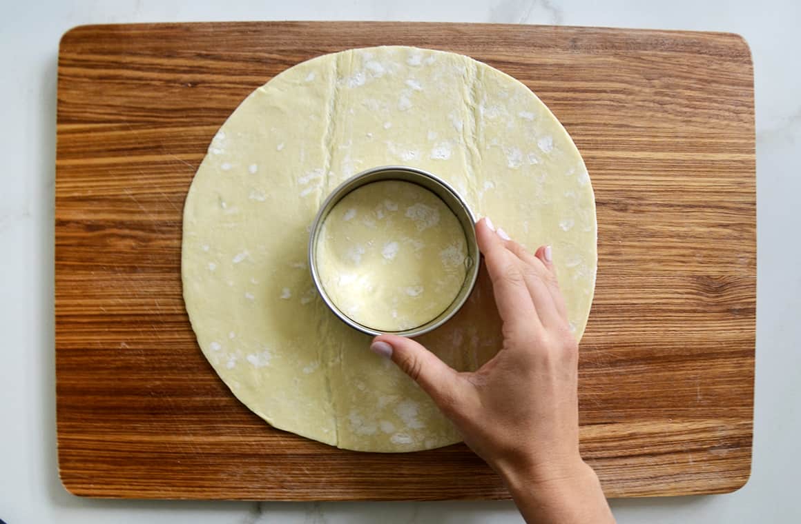 A hand holding a round cookie cutter in the center of a circular piece of puff pastry