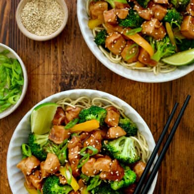 Bowls are filled with chicken and broccoli stir fry, with dishes of sesame seeds and scallions nearby.