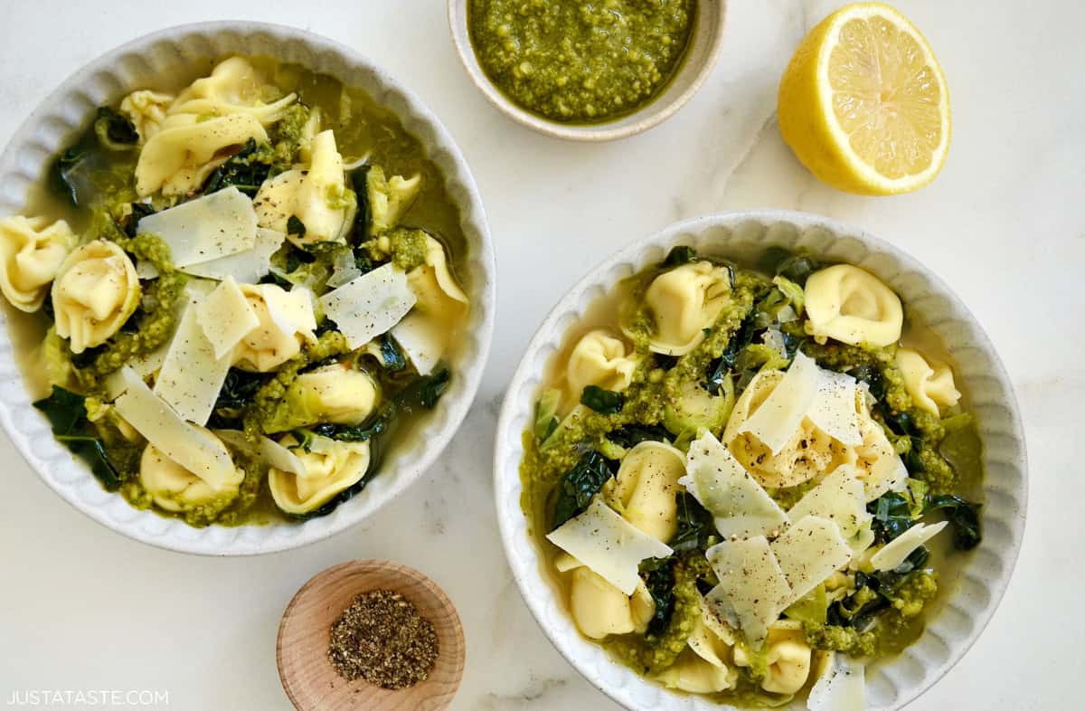 Two bowls of tortellini soup with kale and Brussels sprouts, topped with shaved Parmesan and a swirl of pesto.