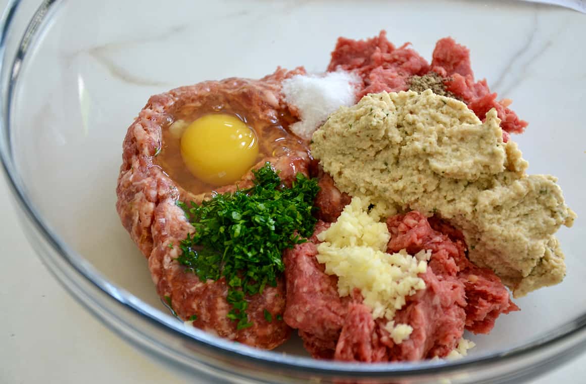 A clear bowl containing ground meat, minced garlic, breadcrumbs, salt, an egg and fresh herbs