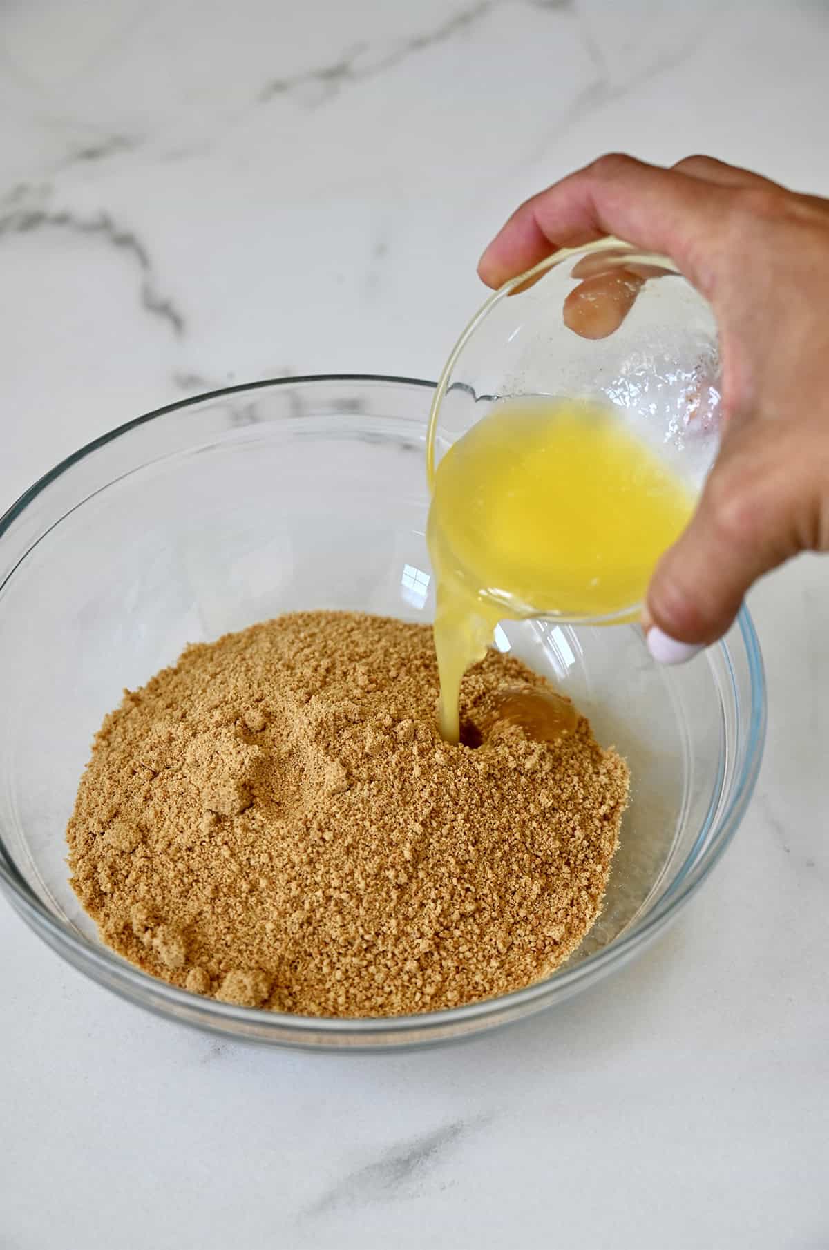 Pouring melted butter into a mixing bowl with finely ground graham cracker crumbs.