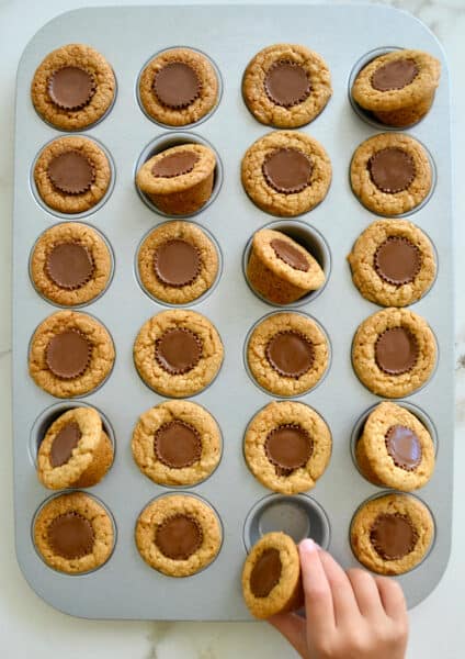 A child's hand reaching for a Mini Peanut Butter Cup Cookie in a muffin tin