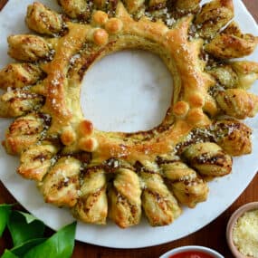 A top-down view of a pull-apart Pesto Puff Pastry Wreath next to fresh basil and small bowls containing marinara sauce and parmesan