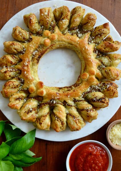 A top-down view of a pull-apart Pesto Puff Pastry Wreath next to fresh basil and small bowls containing marinara sauce and parmesan