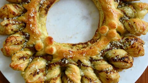 A top-down view of a pull-apart Pesto Puff Pastry Wreath next to fresh basil and small bowls containing marinara sauce and parmesan
